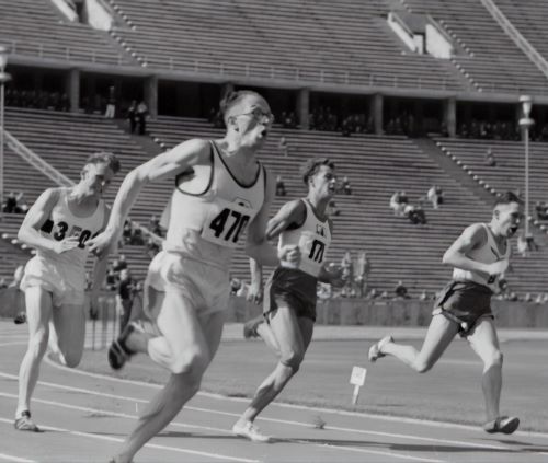 Four athletes sprint on a track in a stadium, wearing numbered jerseys. The stands are mostly empty.