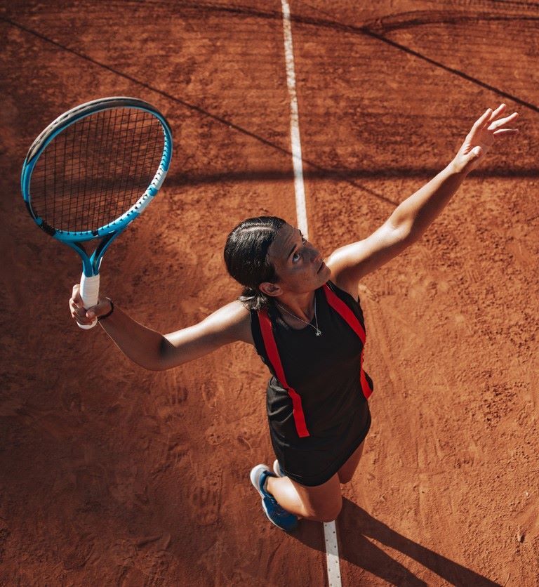 A tennis player in a black outfit is serving a ball on a clay court.