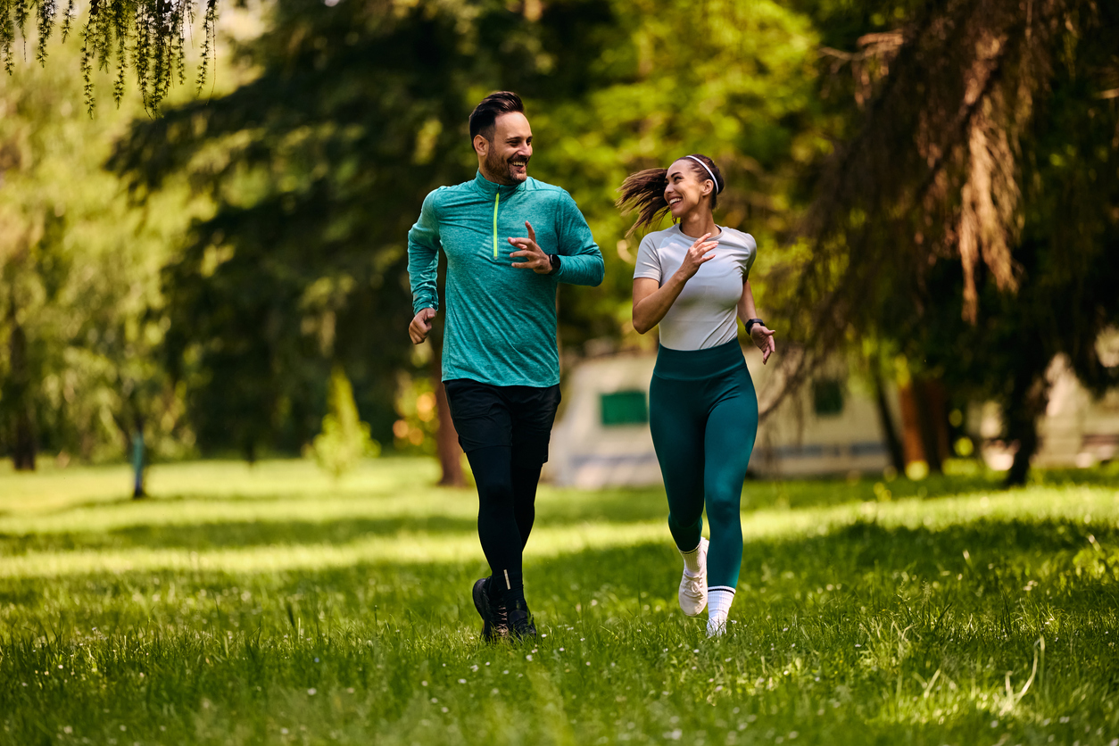A man and a woman are jogging on a grassy field in a park, surrounded by trees. They are both smiling and wearing athletic clothing.